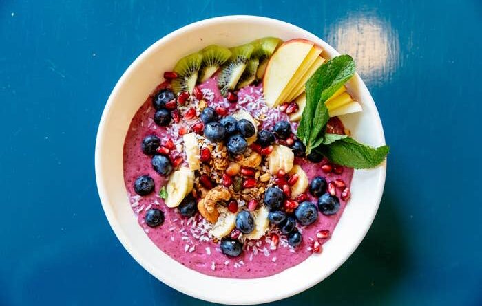 A smoothie bowl topped with blueberries, banana slices, pomegranate seeds, kiwi slices, apple slices, granola, coconut flakes and mint leaves.