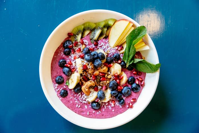 A smoothie bowl topped with blueberries, banana slices, pomegranate seeds, kiwi slices, apple slices, granola, coconut flakes and mint leaves.